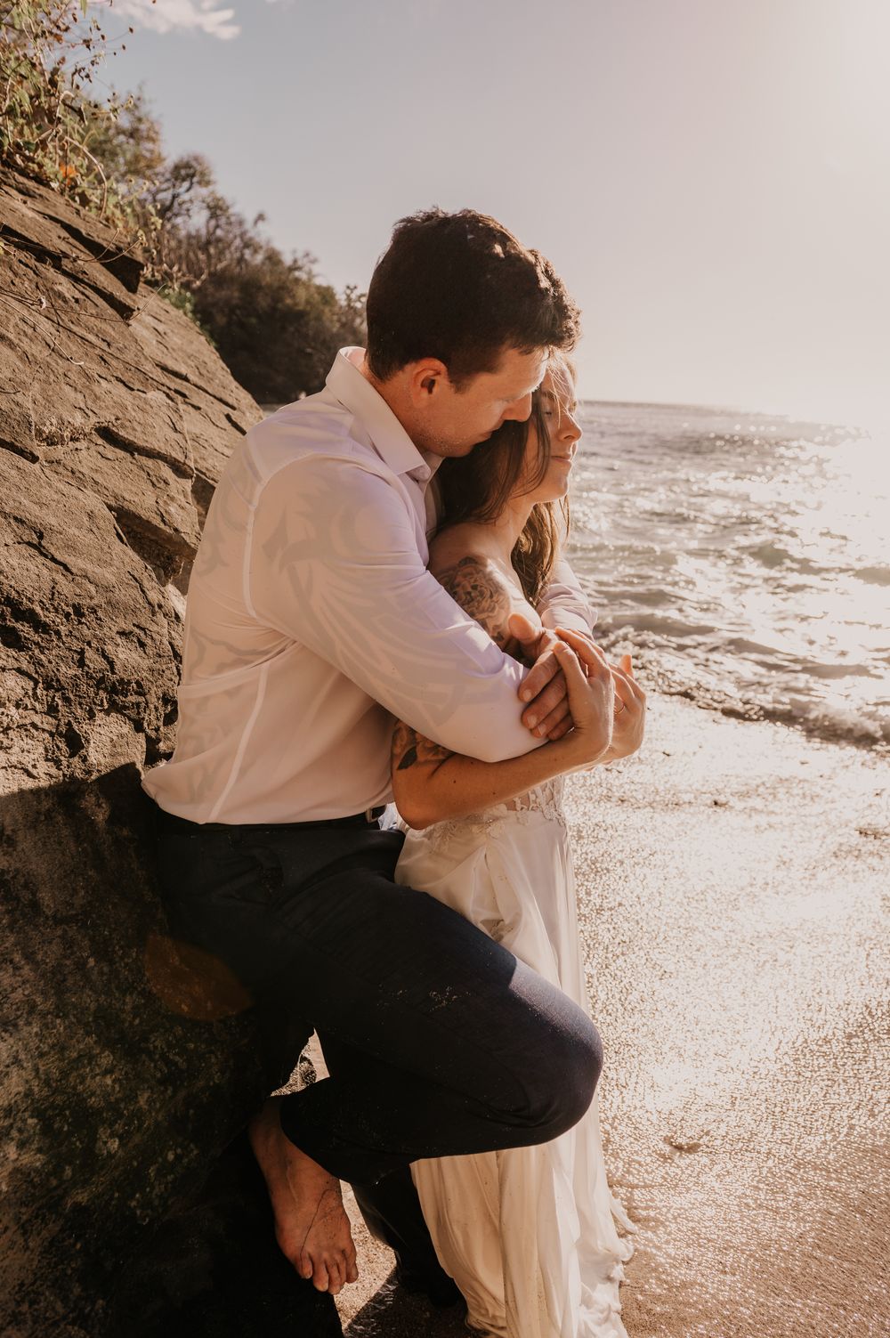 A romantic couple embraces by the rocky coastline at sunset, with waves crashing on the beach in the background.