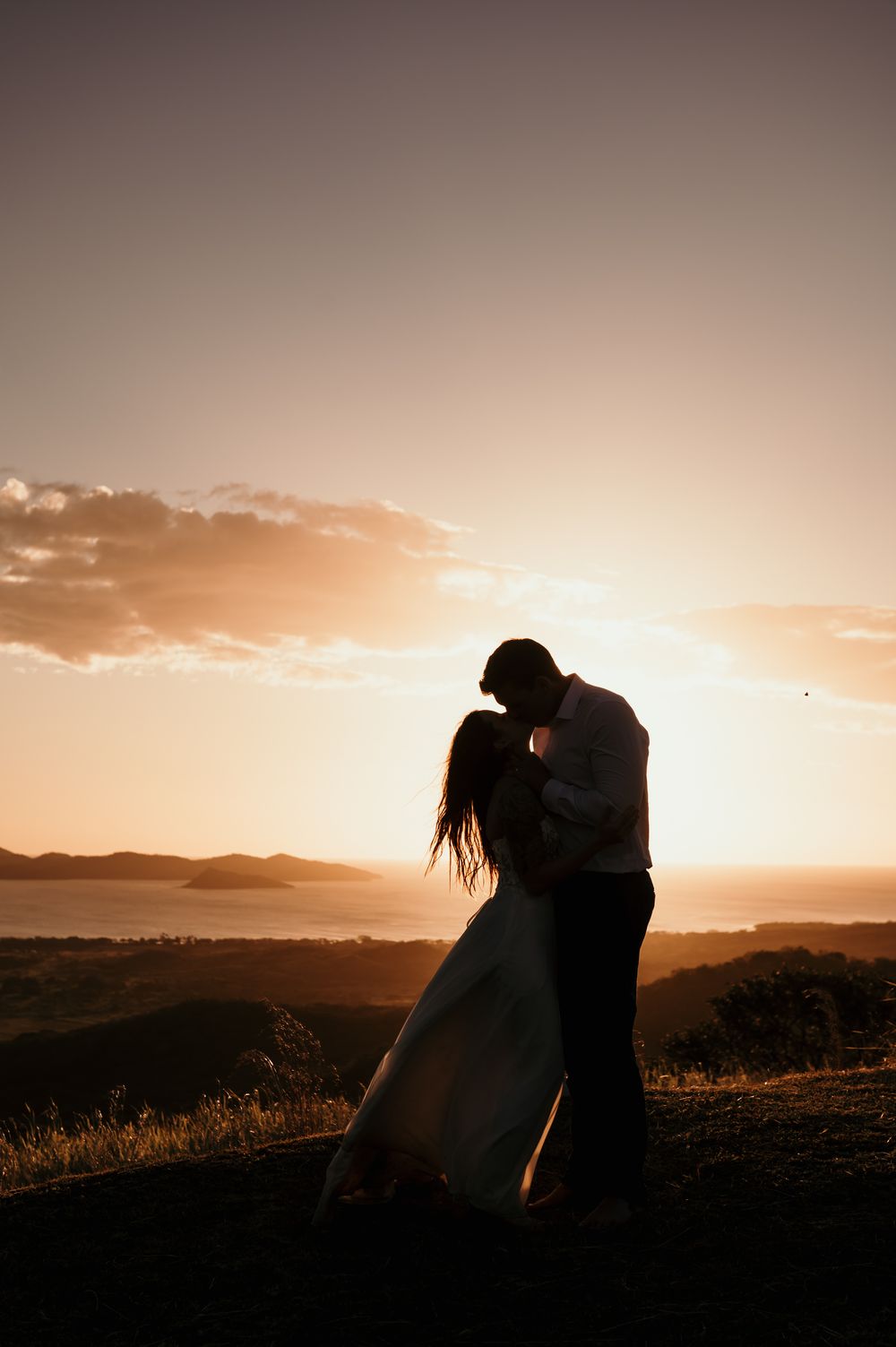 A romantic silhouette of a couple embracing on a hilltop against a golden sunset and mountain landscape.