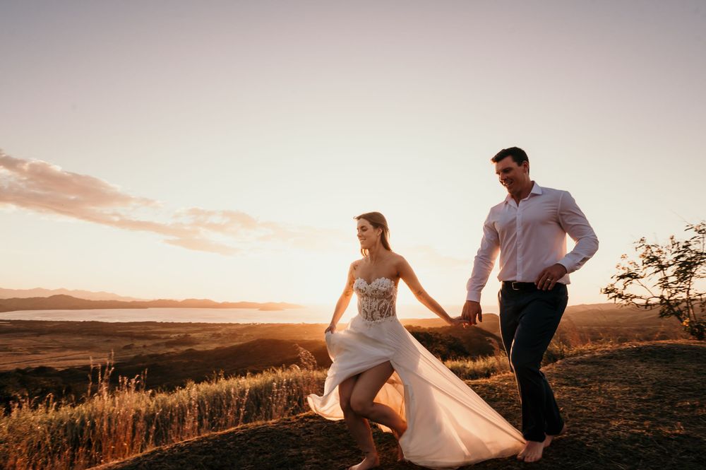 A couple in wedding attire runs together across a hilltop at sunset, silhouetted against a golden sky.