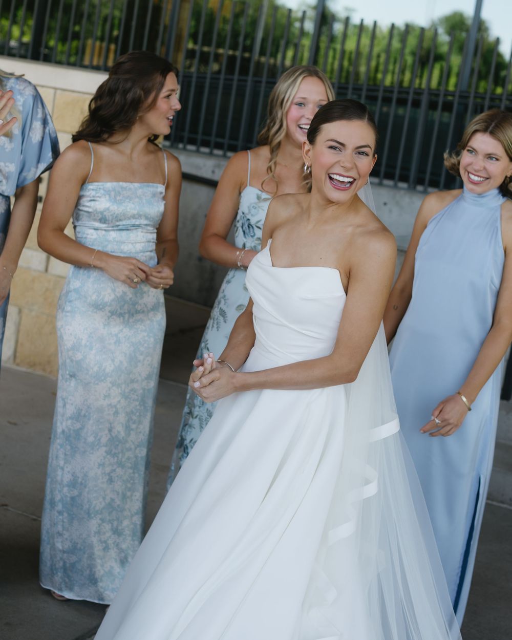 Wedding party laughs joyfully as they pose together, with bridesmaids in pastel dresses surrounding a bride in white.
