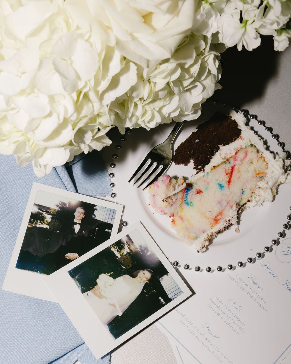 Wedding detail photo showing white carnations, a slice of cake on a plate with a fork, and Polaroid photos.