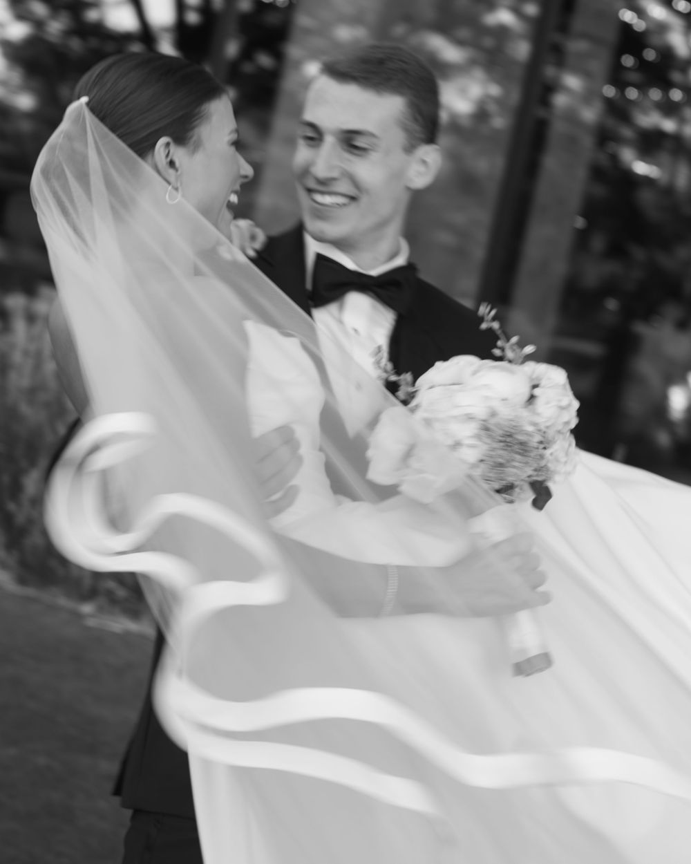 A black and white photo of newlyweds sharing a joyful moment as the bride's veil flows in the breeze.