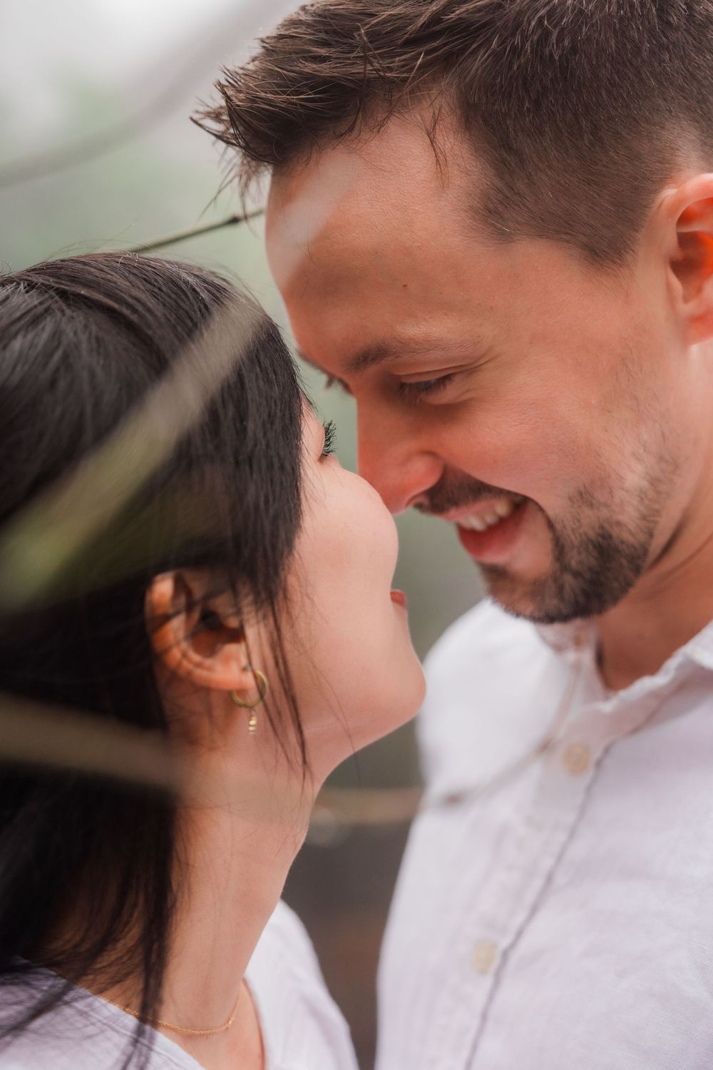 Close romantic portrait of a couple looking at each other with gentle smiles through leaves in soft lighting.