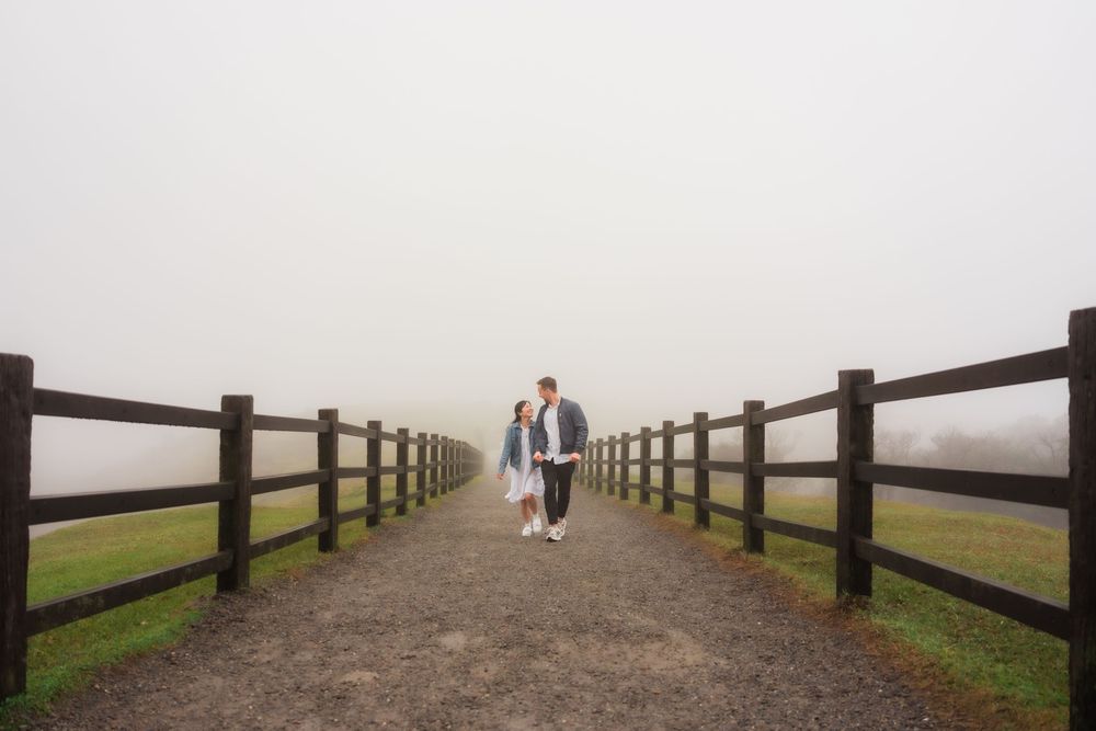 Two people walk hand in hand along a foggy wooden fence-lined path through a misty meadow landscape.