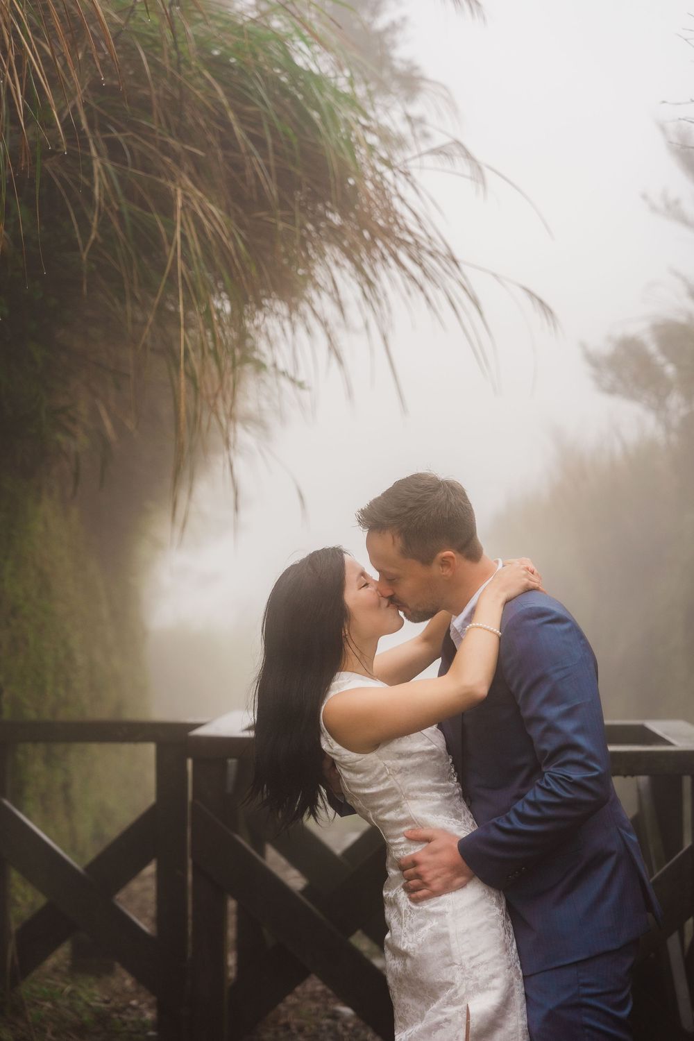 Romantic couple shares an intimate moment on a misty deck with tall grass and foggy scenery in the background.
