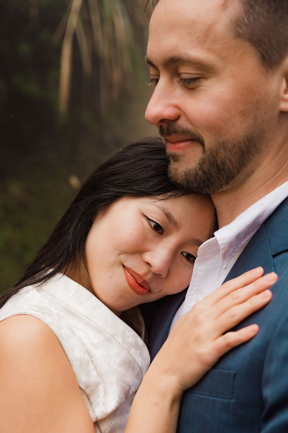 Intimate portrait of a couple embracing outdoors, creating a tender romantic moment at dusk, wearing a white qipao and blue suit.