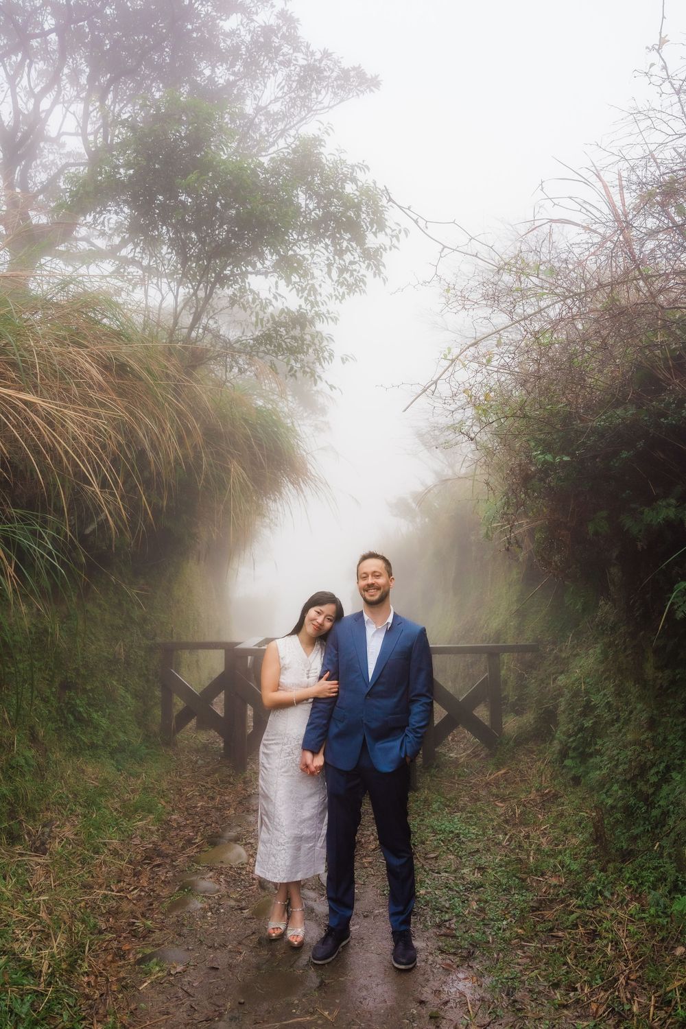 A couple stands on a misty forest path surrounded by tall grass and trees on a foggy day wearing a white qipao and blue suit.