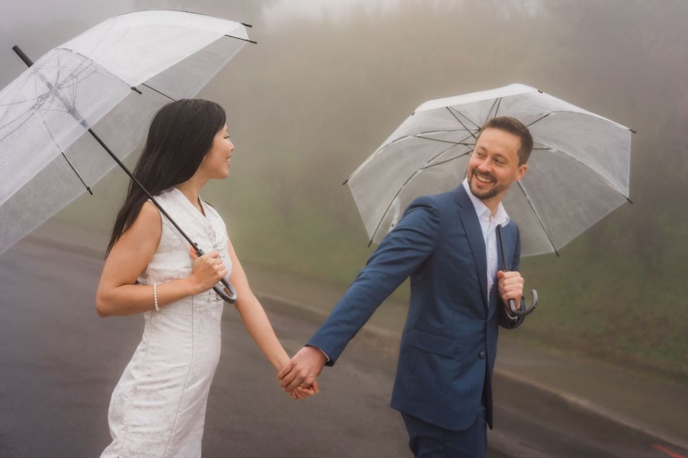 Wedding couple holding hands while walking with clear umbrellas on a misty road.