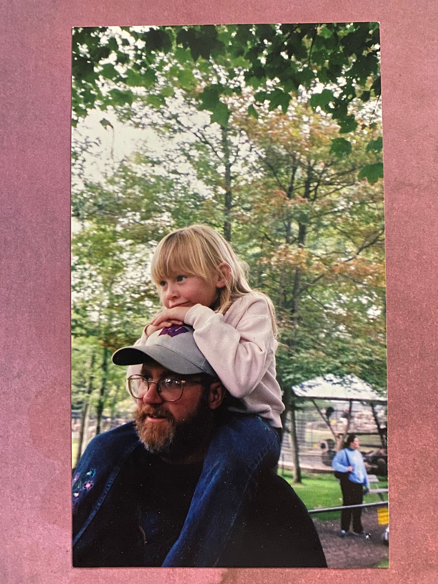A child with blonde hair sits on shoulders in a park while trees and picnic tables are visible in the background.