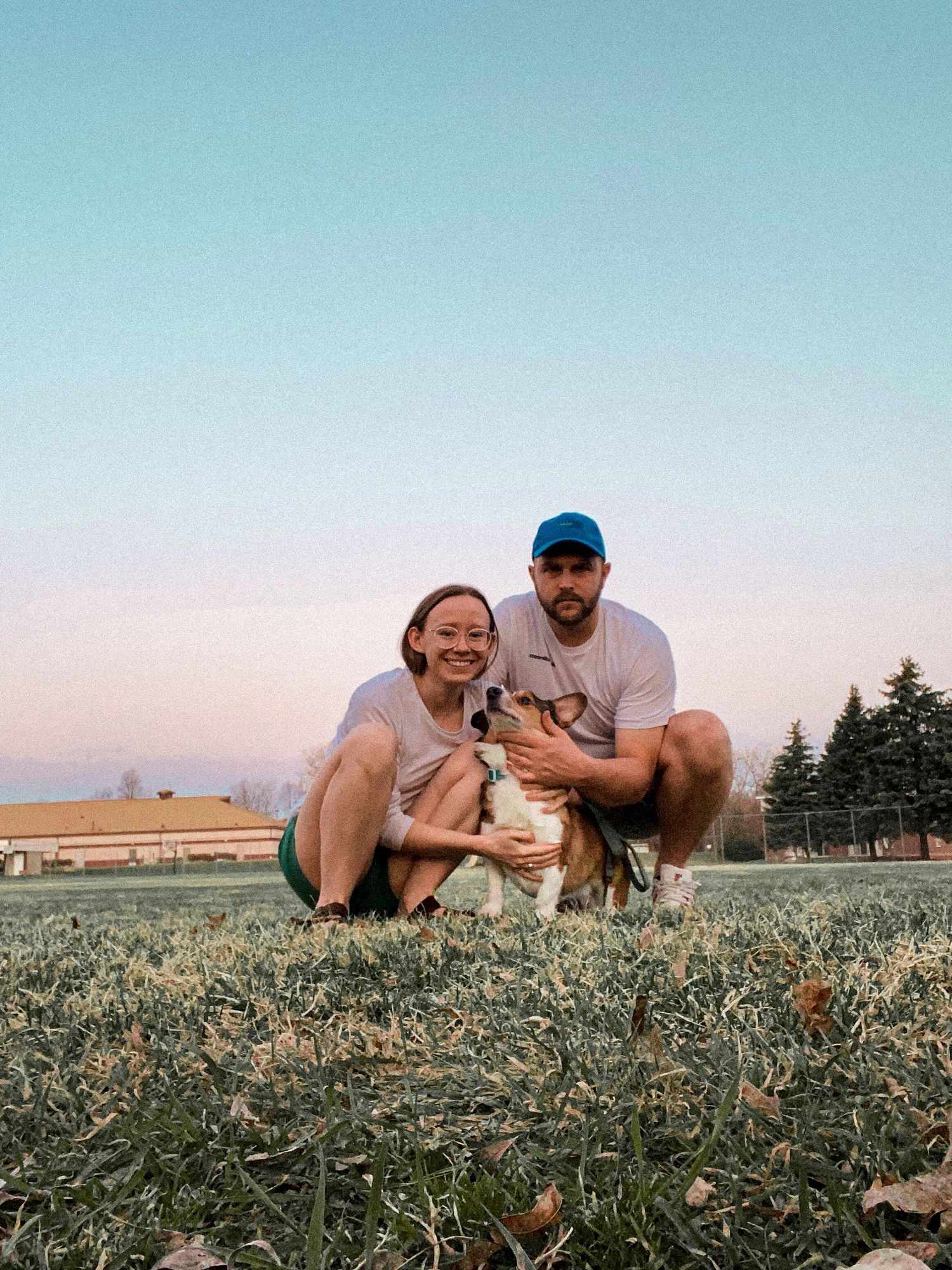 Two people crouching with their dog on a grassy field during sunset with evergreen trees and pink sky in background.