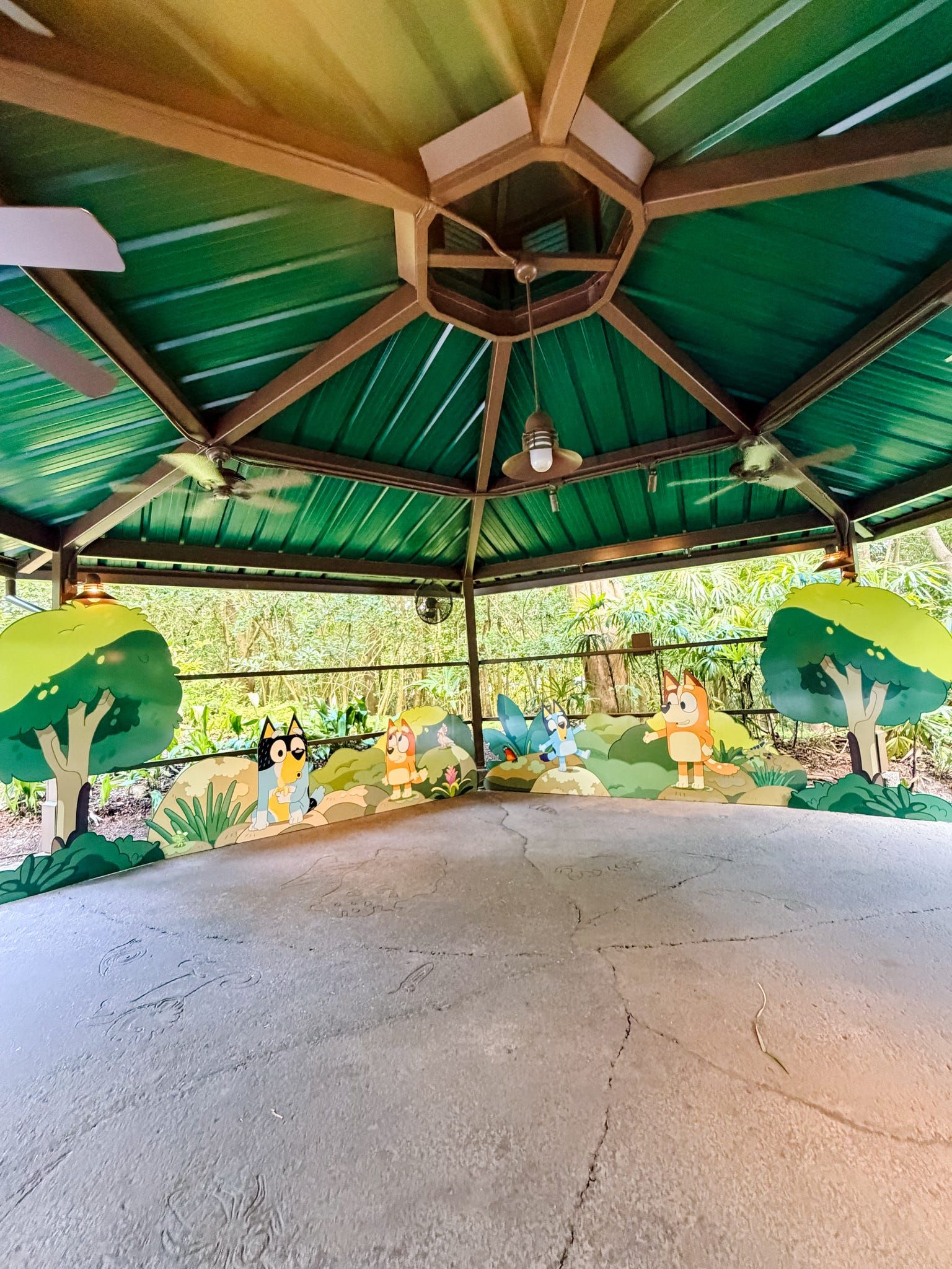 A gazebo with green roof and Bluey cartoon character cutouts decorating the interior walls, surrounded by lush tropical greenery.