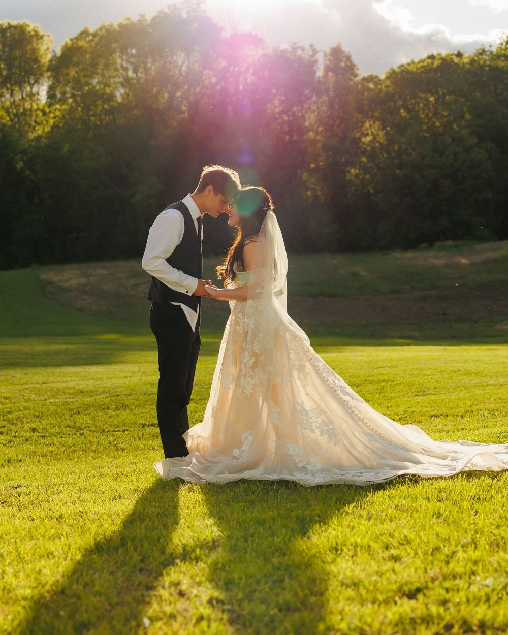 A bride and groom embrace in golden sunlight on a manicured lawn with forest backdrop.