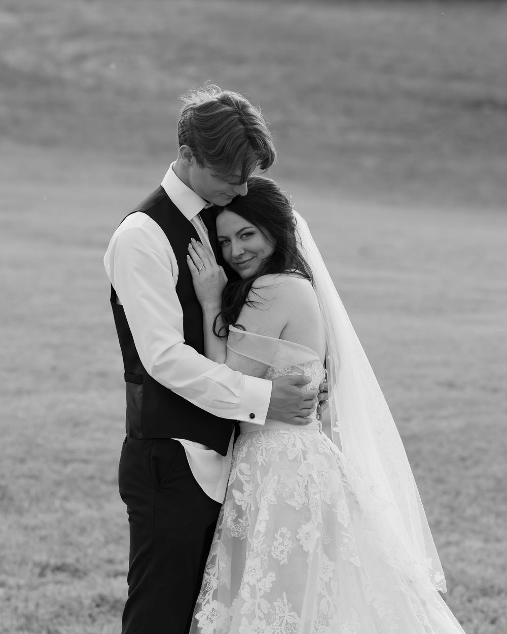 A tender black and white wedding photograph showing a couple embracing outdoors in formal attire with a flowing veil.