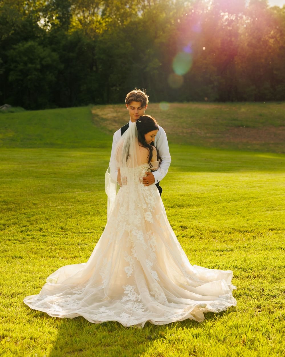 A couple stands embracing on a sunlit grassy field at golden hour, with the bride wearing a flowing lace wedding gown.
