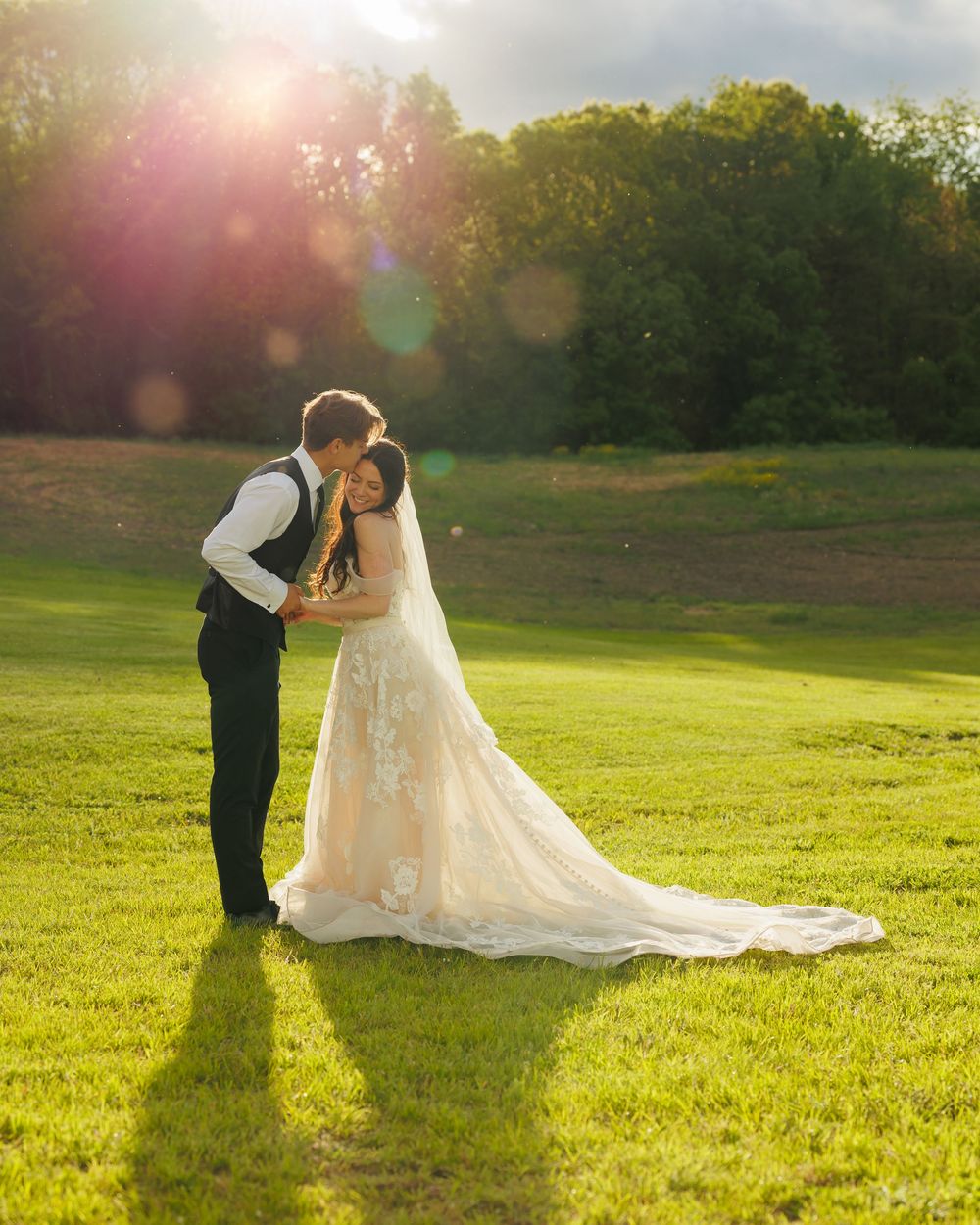 A romantic wedding couple shares an intimate moment at sunset on green grass with a tree-lined backdrop.