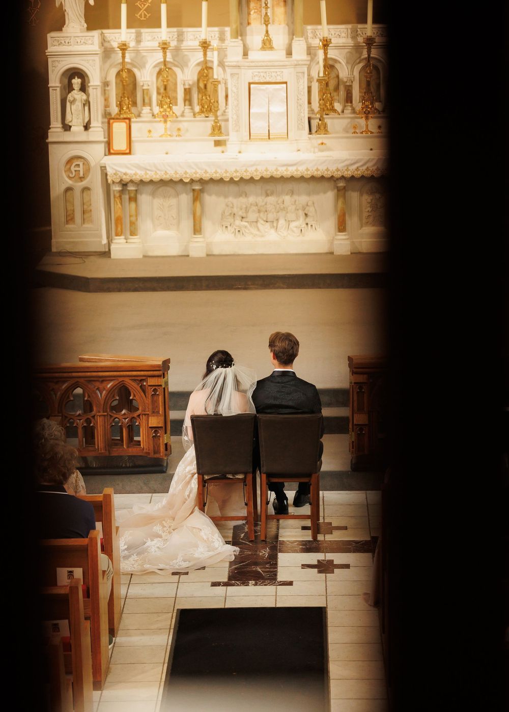 A couple sits together on a wooden church pew facing an ornate altar in a dimly lit sanctuary.