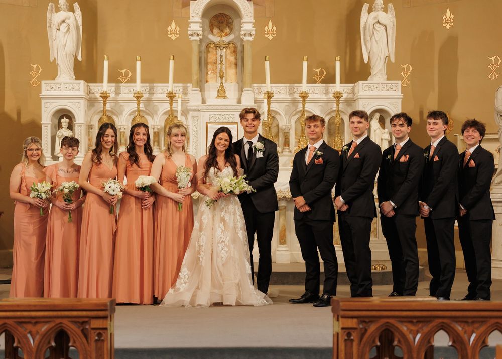 Wedding party stands formally posed in front of an ornate church altar with candles and religious statues.