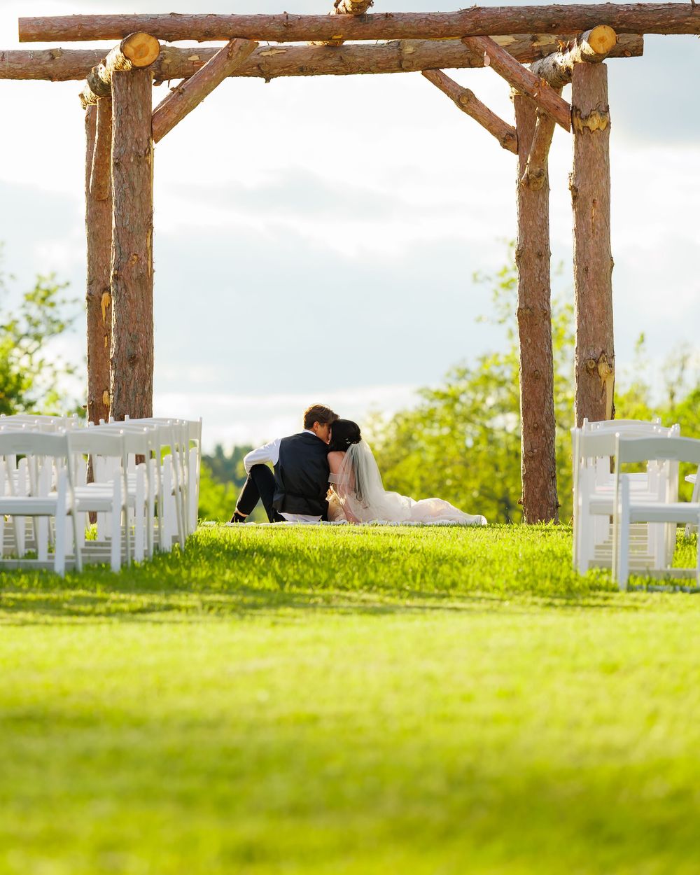 A rustic wooden arbor frames empty white chairs set up for an outdoor wedding ceremony on a grassy hillside.