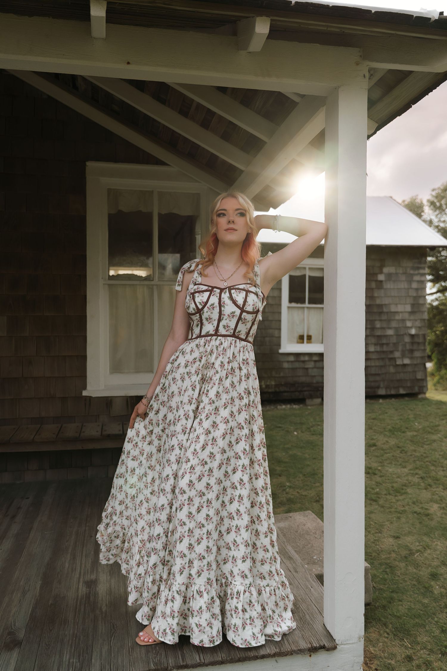 A flowing floral print maxi dress with corset bodice is styled on a rustic farmhouse porch at sunset.