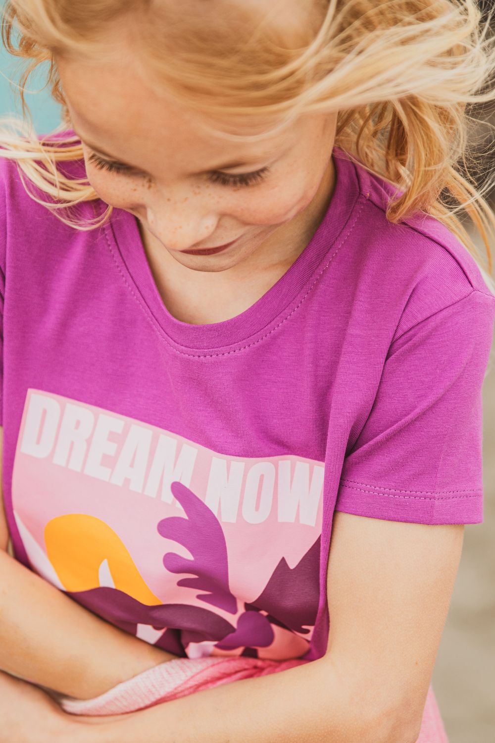 Woman wearing a bright pink t-shirt with graphic design smiling while wind blows through blonde hair.
