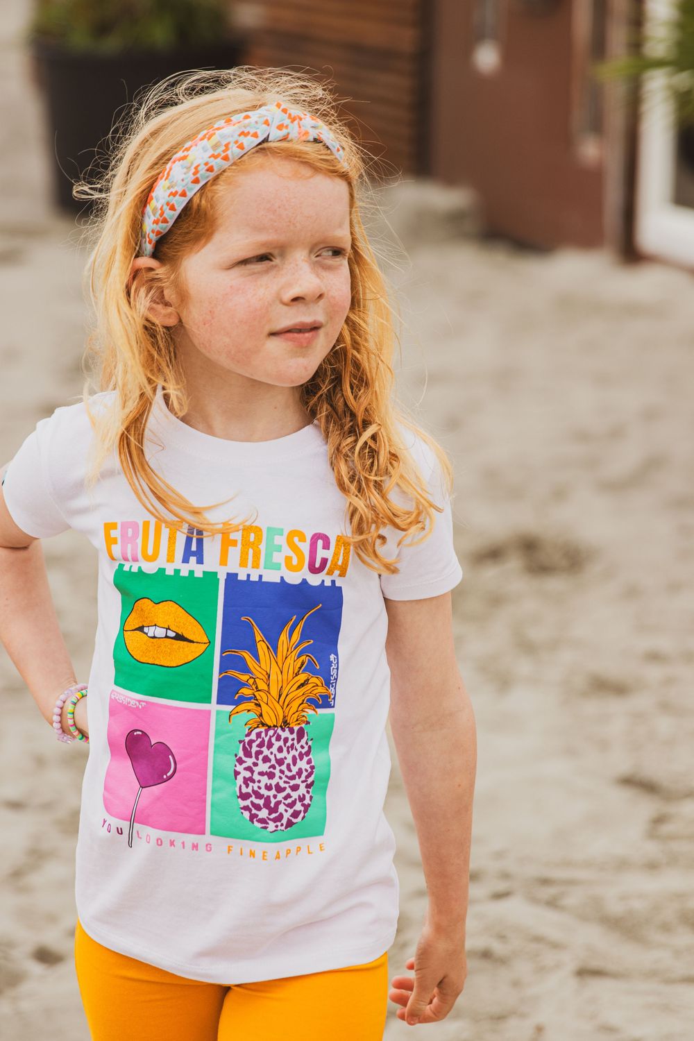 Young redhead wearing a floral headband and colorful graphic t-shirt with fruit designs and orange pants.