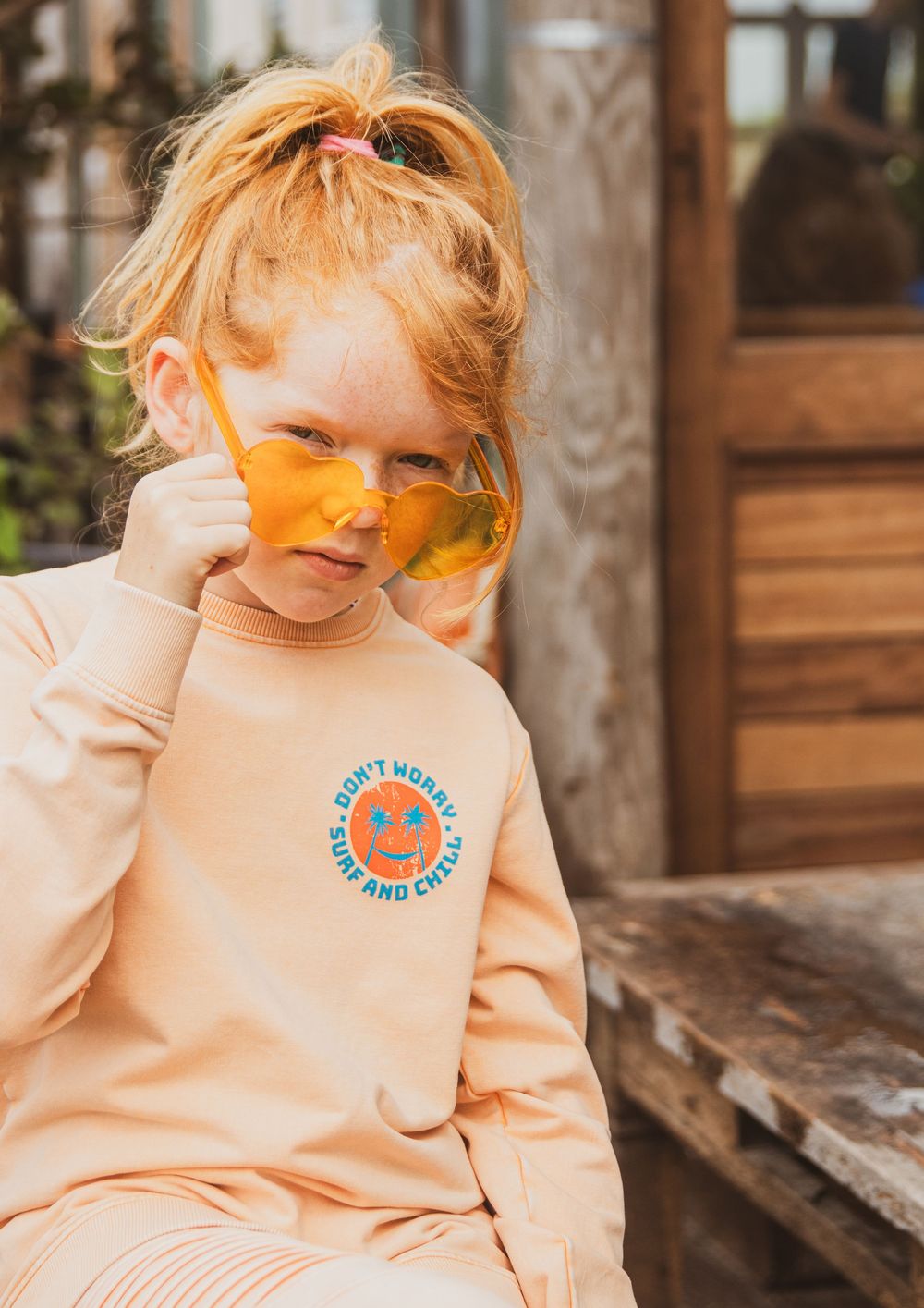 A small child in peach-colored outfit and yellow tinted sunglasses poses on wooden steps outdoors.