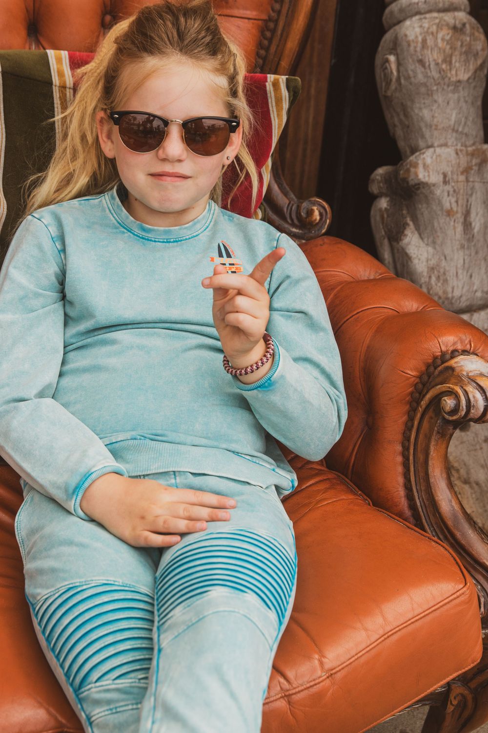 A young person wearing sunglasses and a turquoise sweater relaxes on a brown leather armchair.