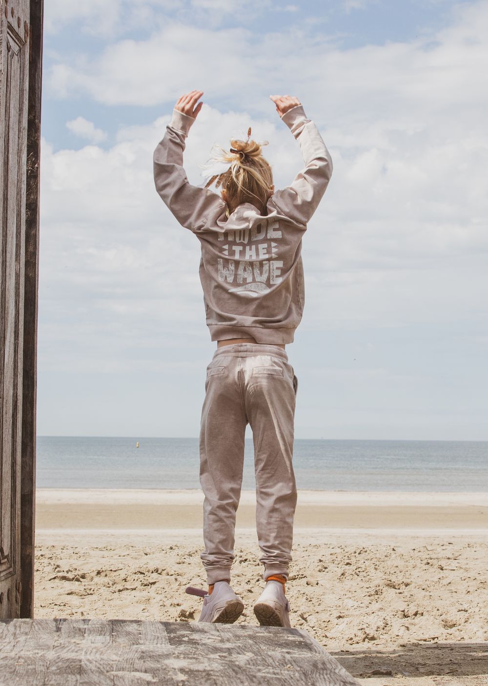 Someone in a beige sweatsuit with 'The Future is True' text stretches their arms up while standing on a beach.