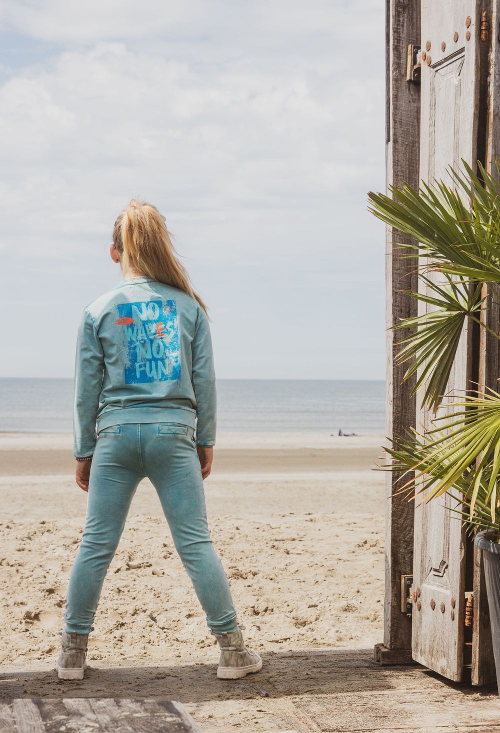 Person in teal denim outfit stands at beach entrance looking towards ocean with palm tree frond visible on right.