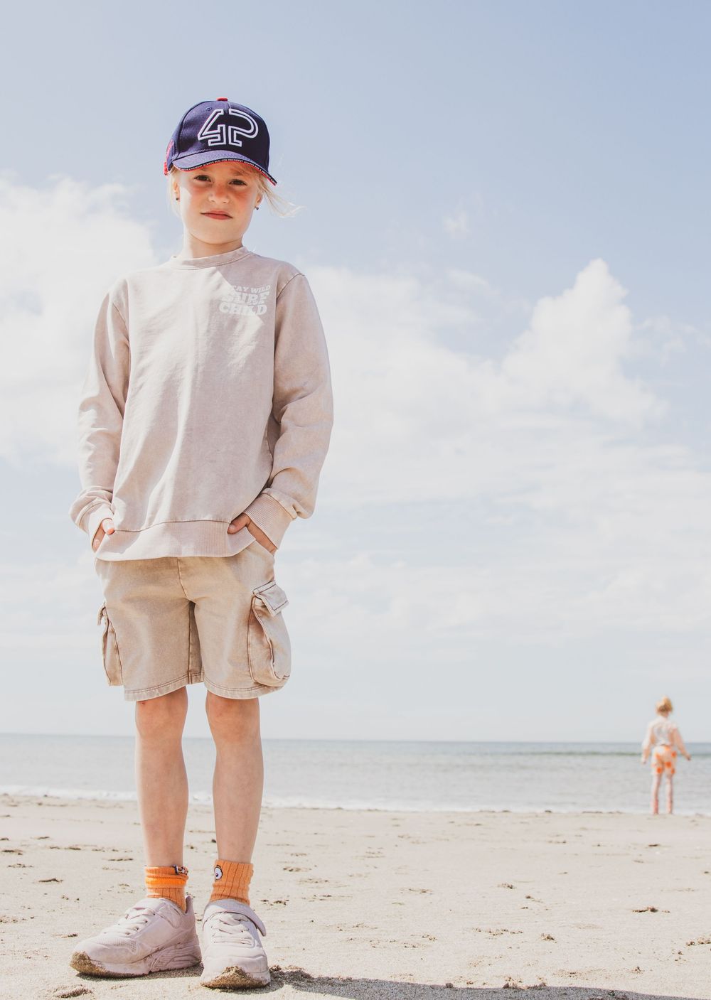 Young person in baseball cap and beige outfit stands casually on a sunny beach with ocean and sky in the background.