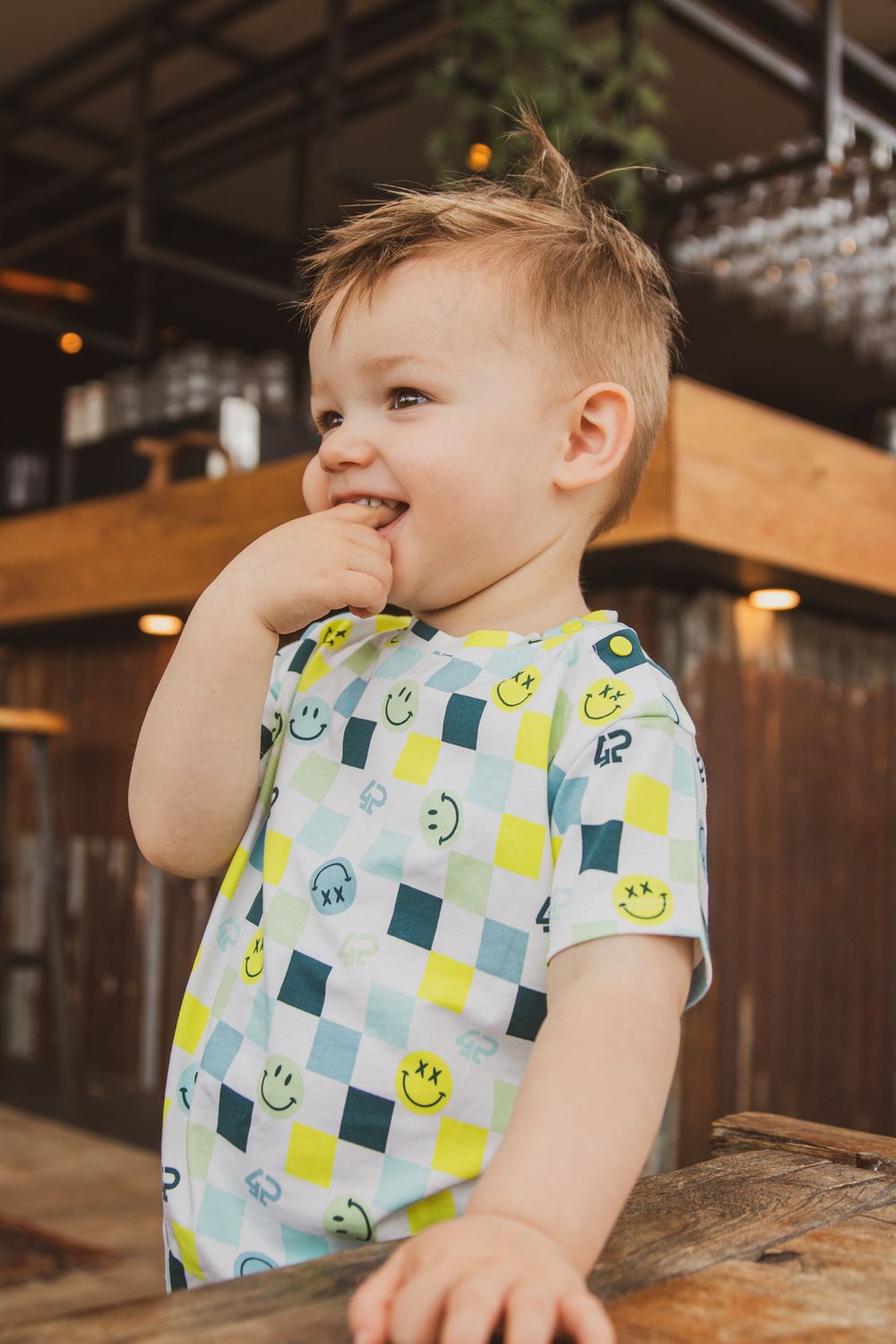 Young child wearing a cheerful blue and yellow smiley face patterned t-shirt laughs while at a restaurant.