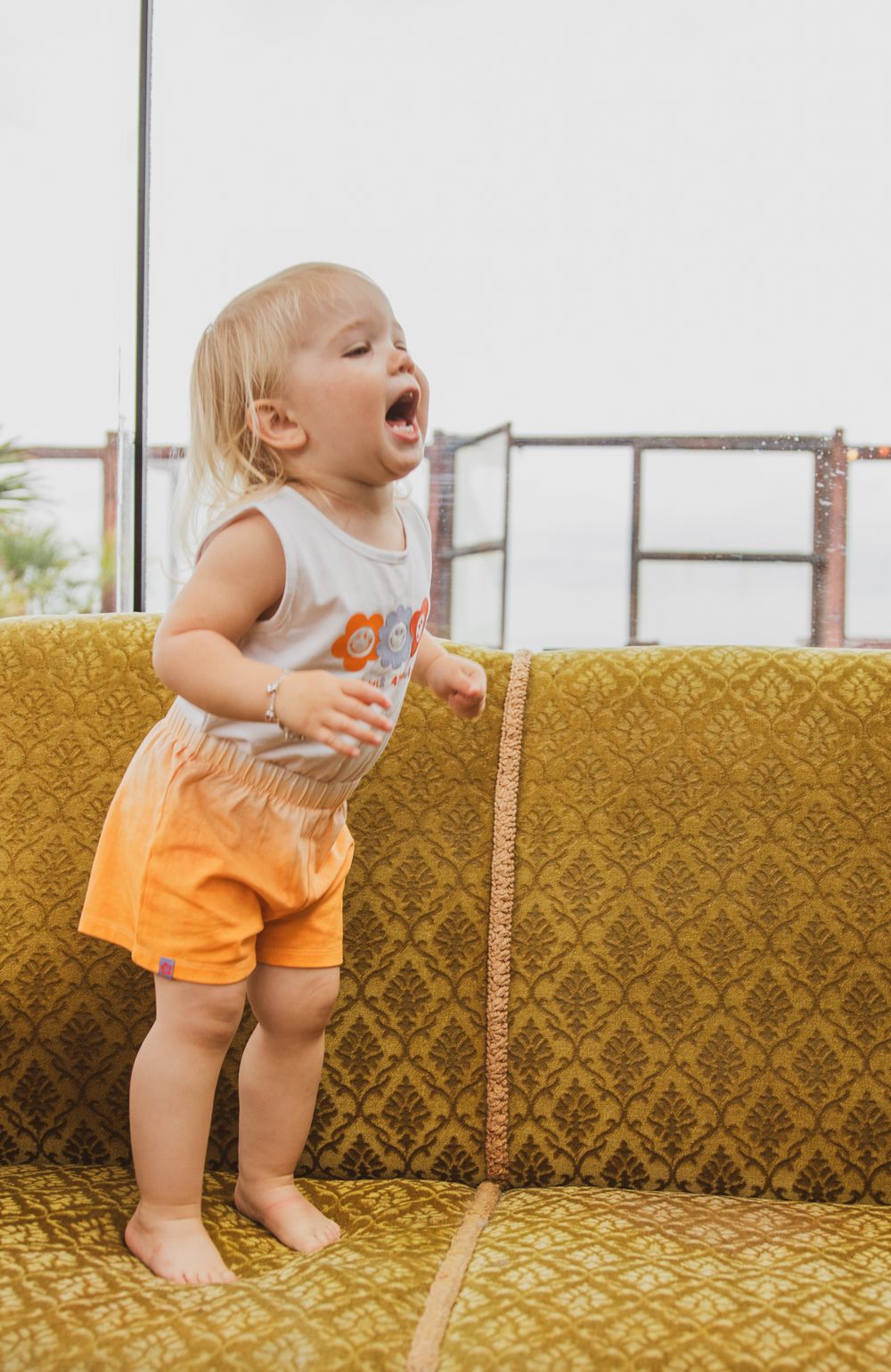 Toddler in orange shorts stands barefoot on yellow vintage couch while playing with a measuring tape.