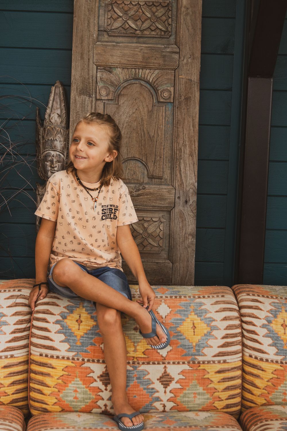 A child sits on a southwestern patterned couch against a rustic wooden door with ornate carvings.