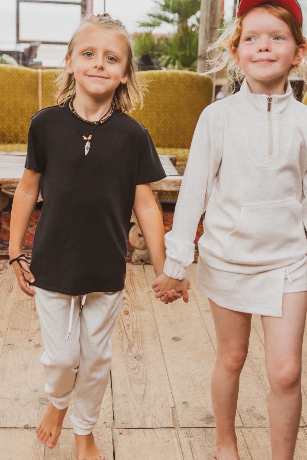 Two children holding hands on a wooden deck wearing casual summer clothing in neutral colors.