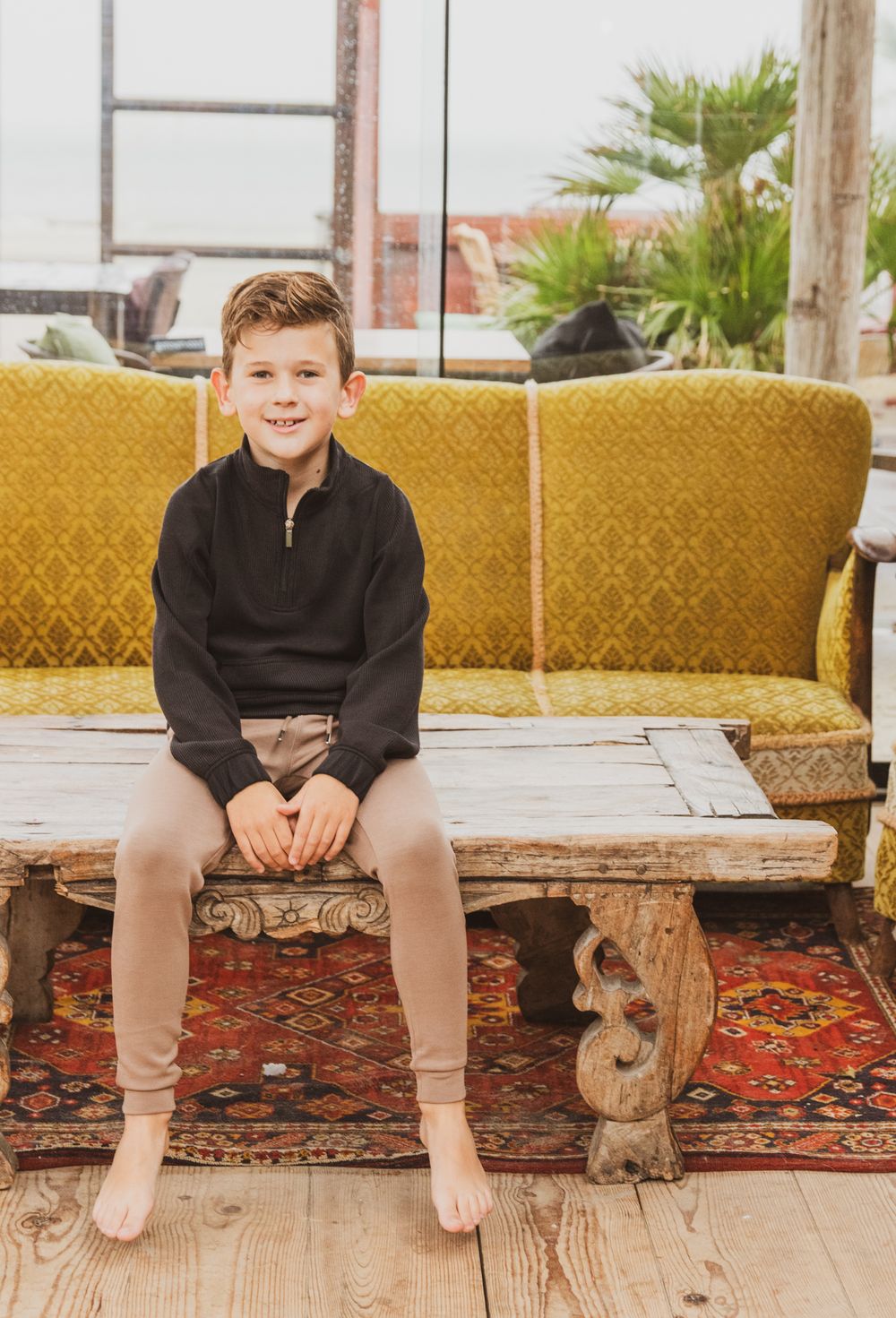 A young figure in a black pullover sits barefoot on a rustic wooden bench with yellow cushions and oriental rug beneath.