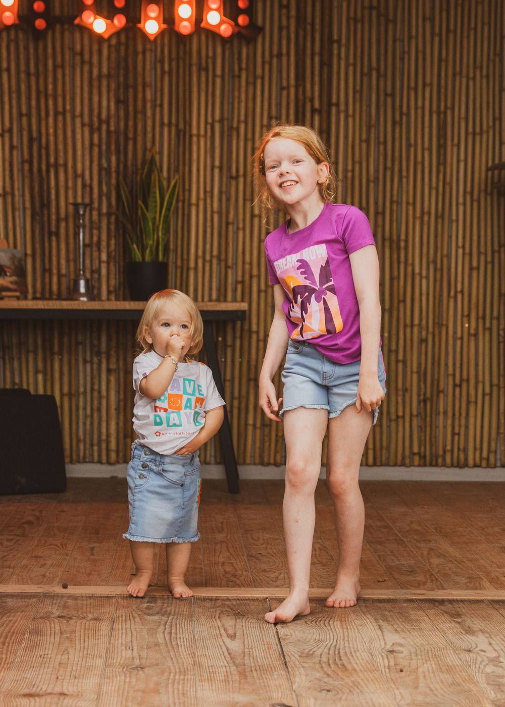 Two children in matching denim shorts and t-shirts stand barefoot against a bamboo wall with red lanterns overhead.