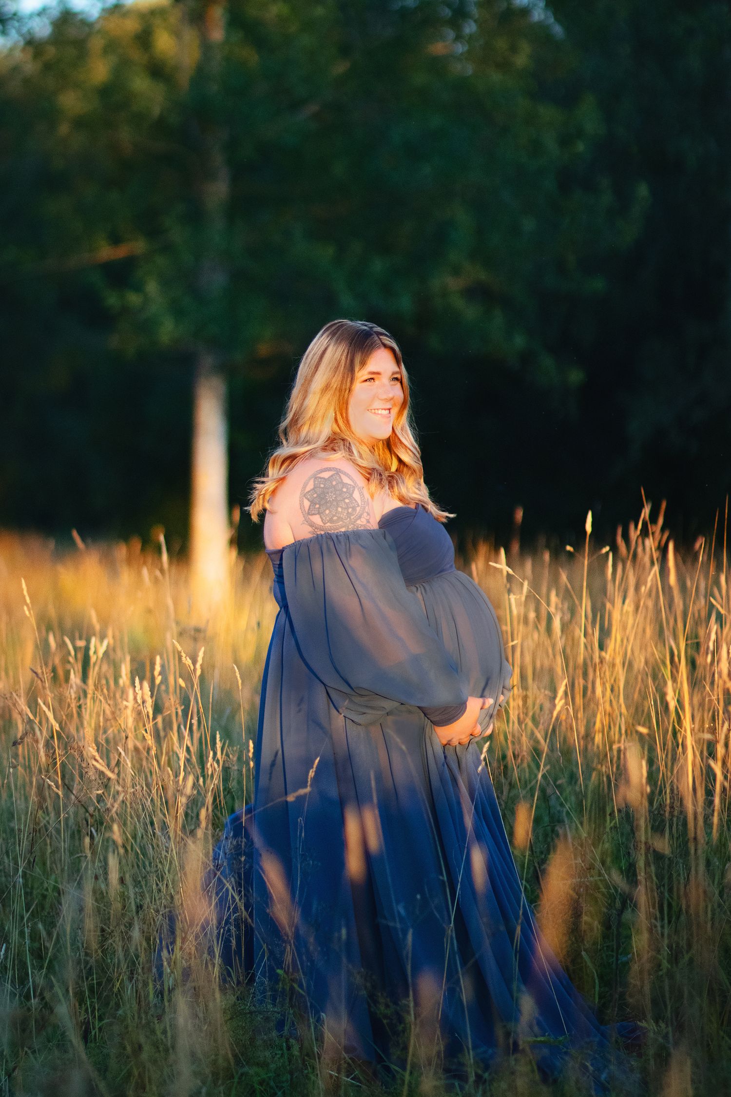 Smiling pregnant woman in sheer blue gown cradling belly in golden sunset field.