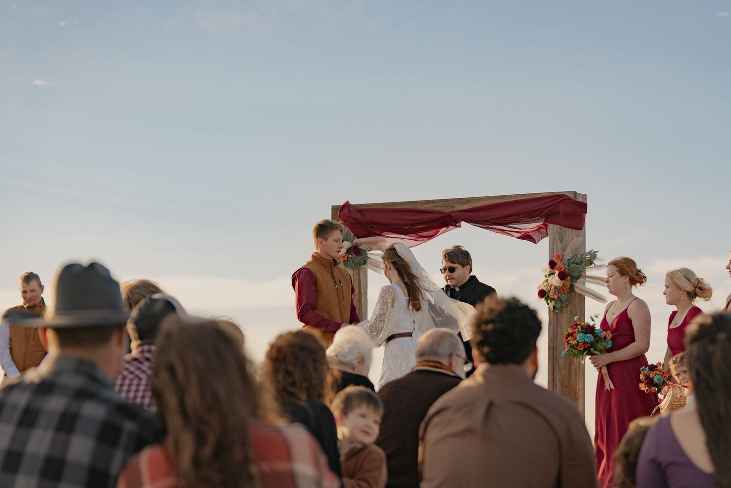 A wedding couple in intimate embrace with bride wearing lace dress and groom in brown vest against burgundy curtain.
