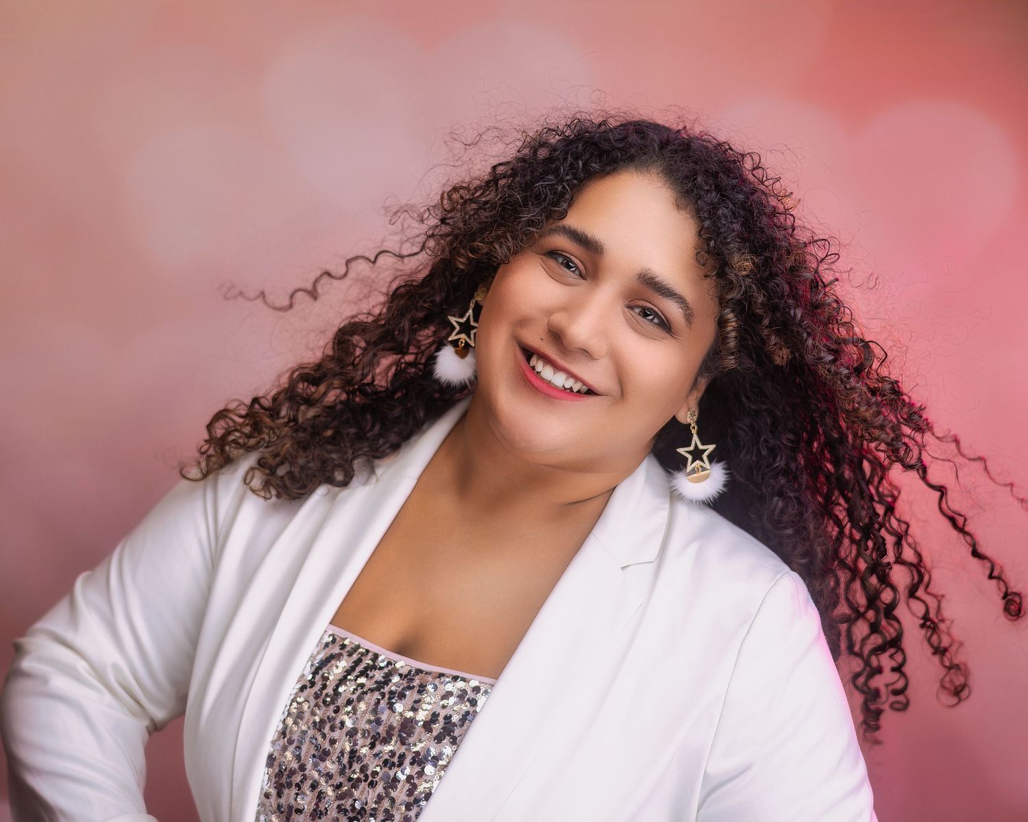 Smiling woman with curly hair in a white blazer and star earrings, standing against a soft pink bokeh background Image Carolina Waldroff