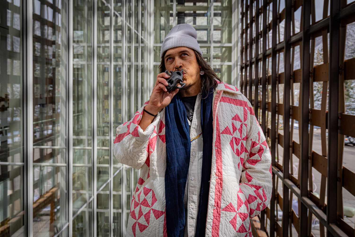 Person wearing a gray beanie and pink patterned coat stands in front of glass blocks while speaking into a microphone.