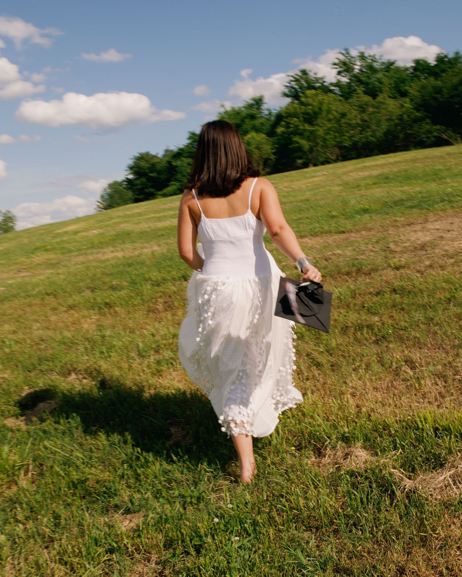Woman in white lace dress walks barefoot through a sunny grassy field holding shoes.