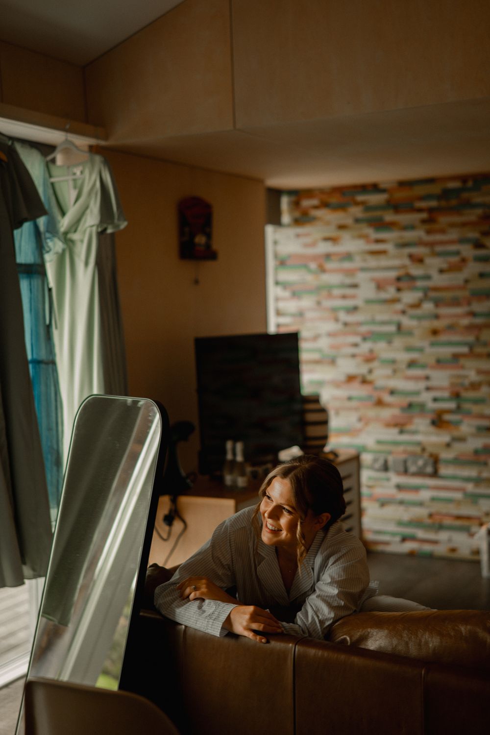 A cozy indoor scene with warm lighting and someone resting on a brown couch near a window with a geometric patterned wall.