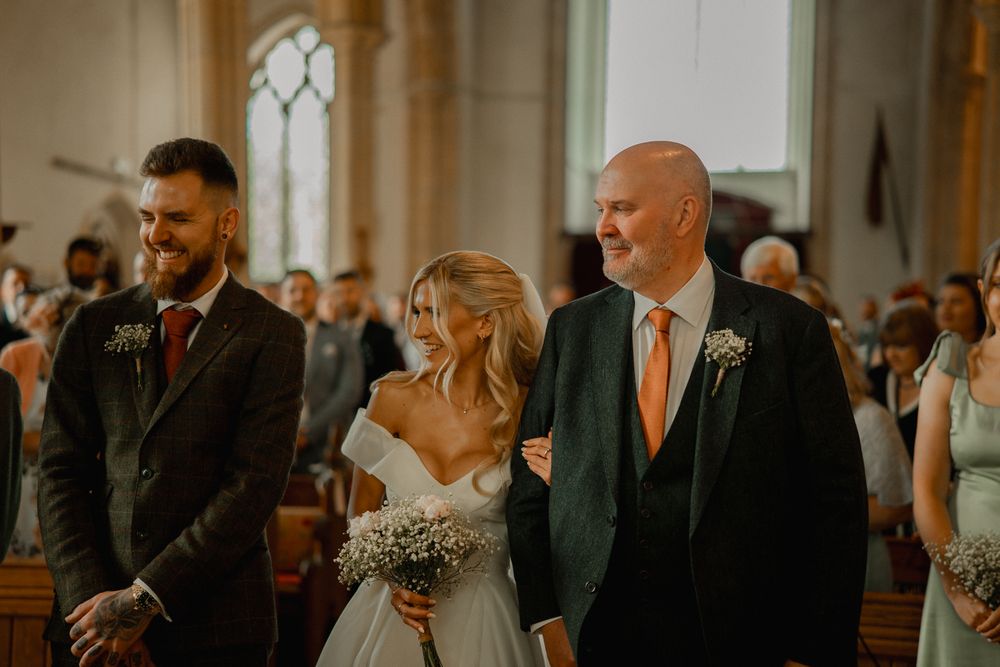 Inside a church during a wedding ceremony, the bride in a white gown walks down the aisle with escort in a dark suit.