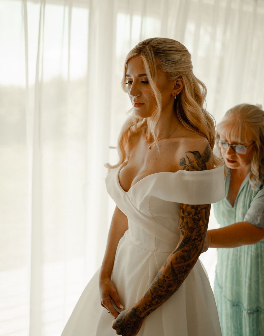 A bride in a white off-shoulder wedding dress gets help with final preparations in a sunlit room.
