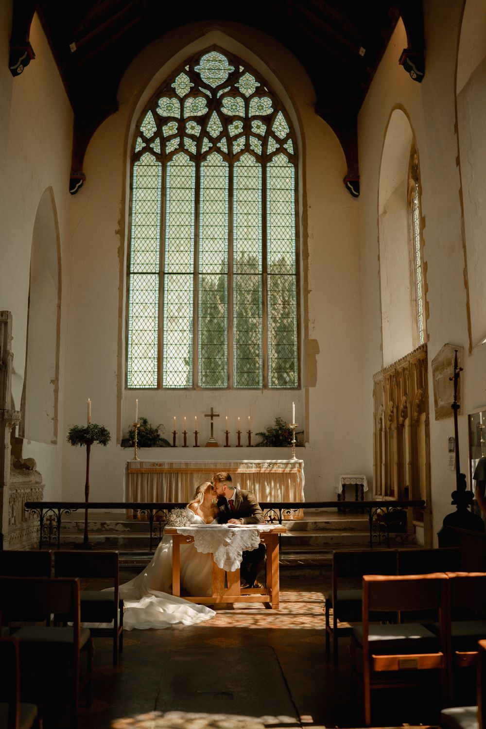 Couple signing marriage documents at altar beneath large stained glass window in historic church.