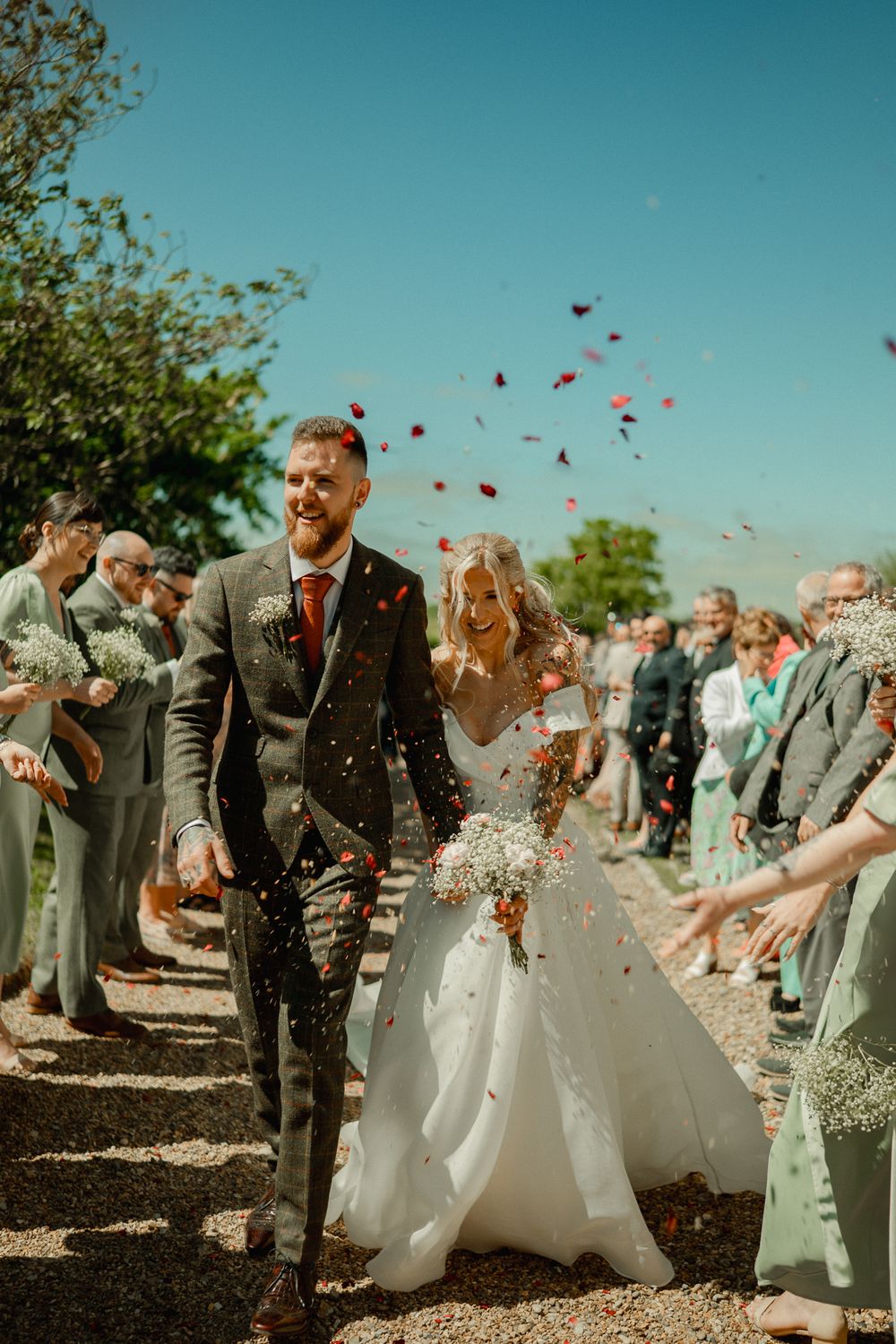 Newlyweds walk down path while guests throw confetti on sunny wedding day.