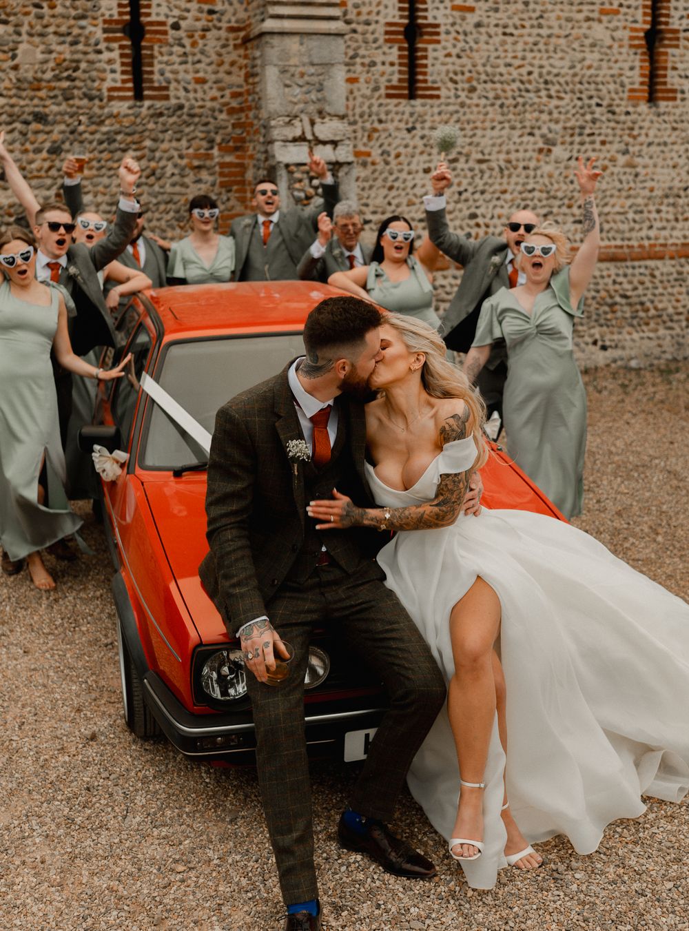 A newlywed couple shares a kiss on a vintage red car while wedding party celebrates in the background.