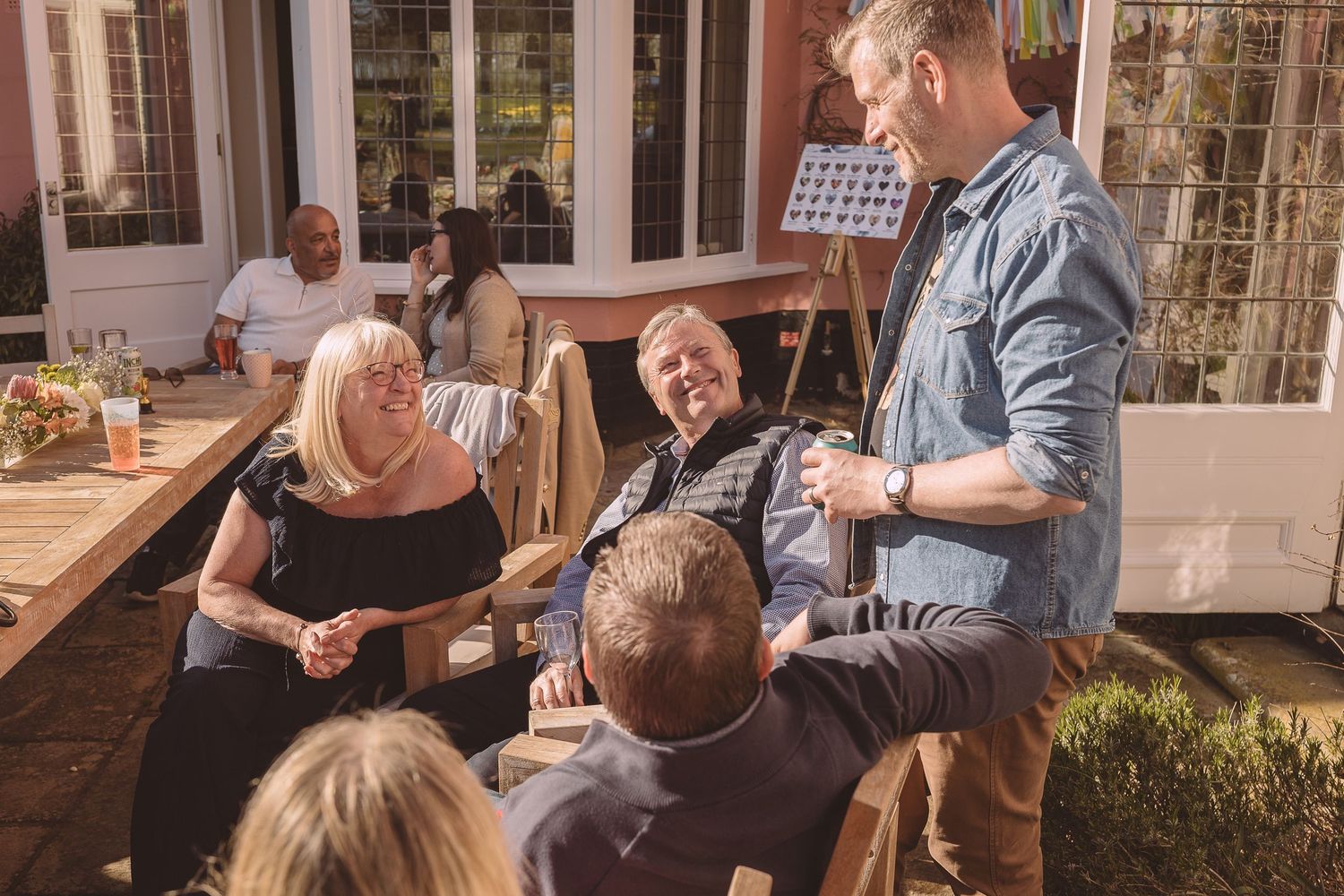 Friends enjoy an outdoor gathering around a wooden table on a sunny patio during daytime.