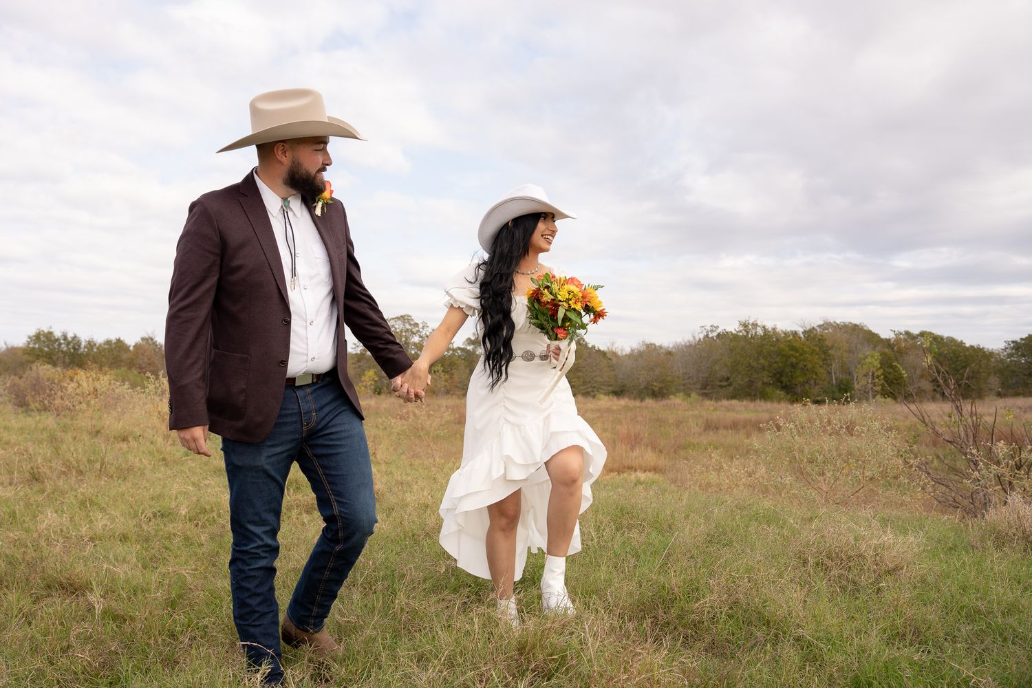 A couple walks hand in hand through a rustic field, with the bride holding yellow sunflowers and wearing a flowing white dress.