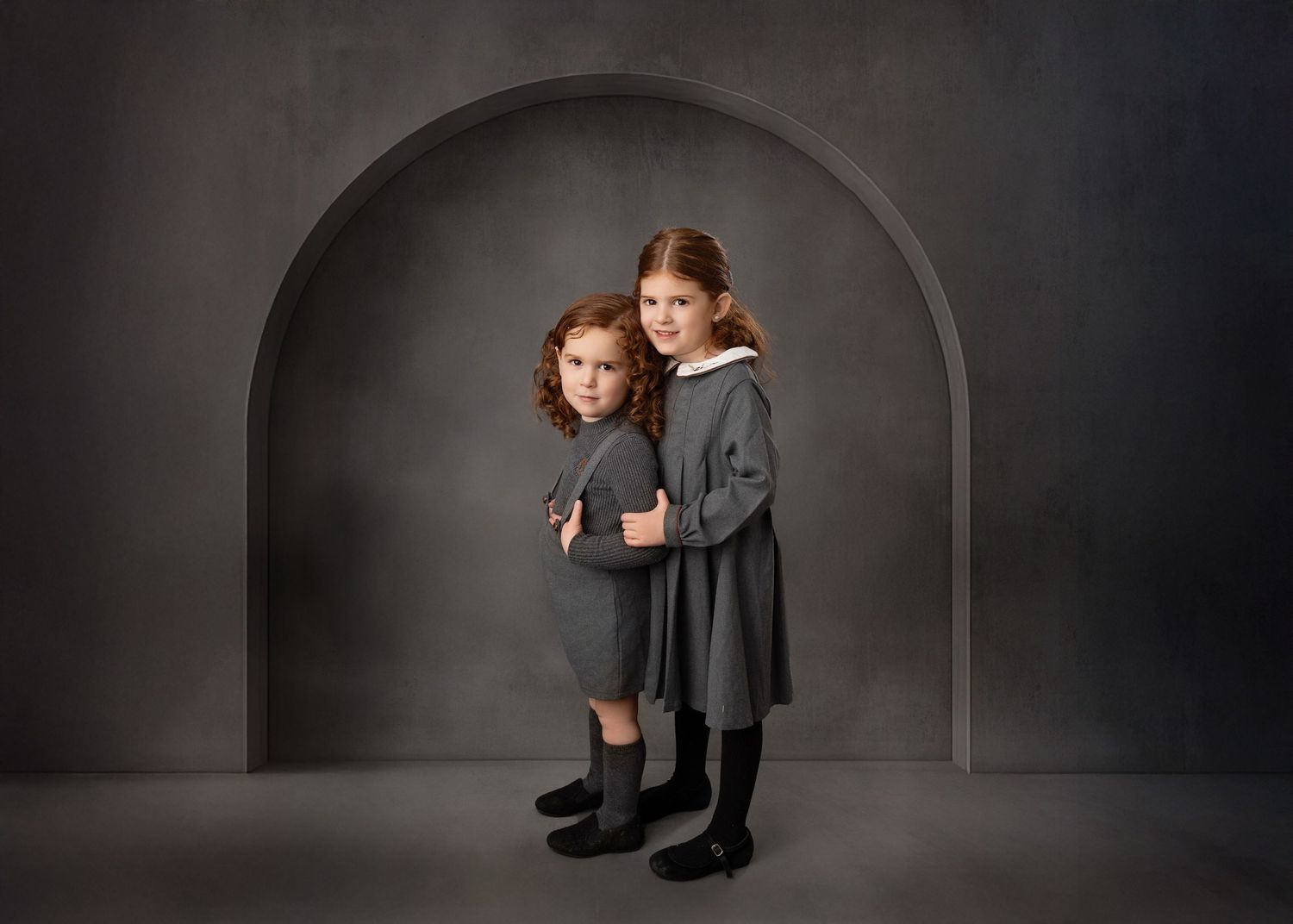 Two children pose in vintage-style clothing against a dramatic arched dark backdrop.